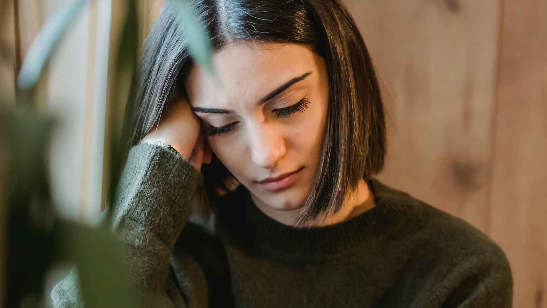 Weary female with closed eyes in casual clothes leaning on hand sitting near window