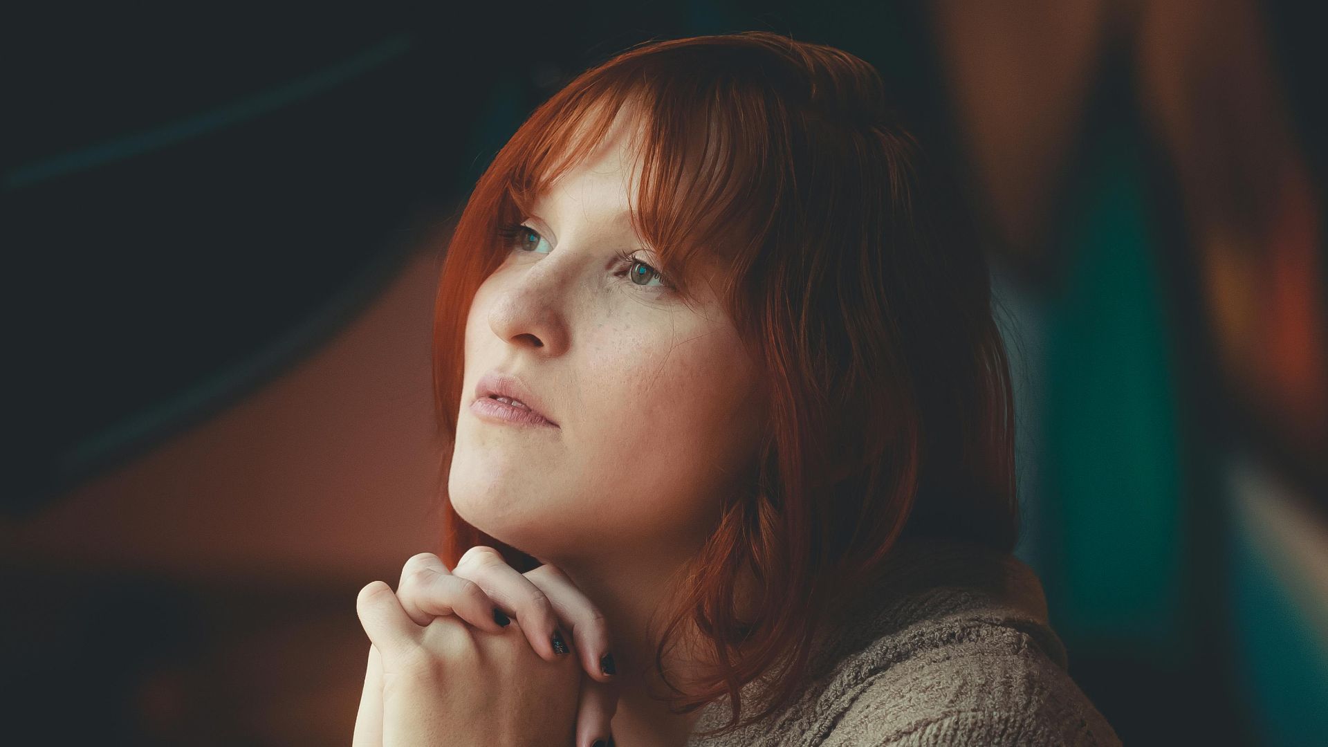 Portrait of a ginger-haired woman sitting thoughtfully, hands clasped at a table indoors.