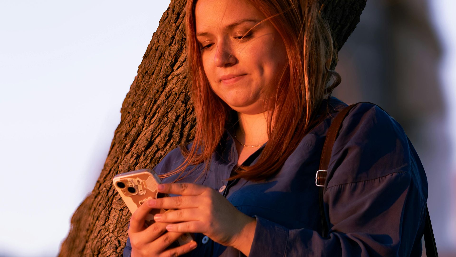 Young woman using smartphone leaning against tree during sunset in Assos, Türkiye.