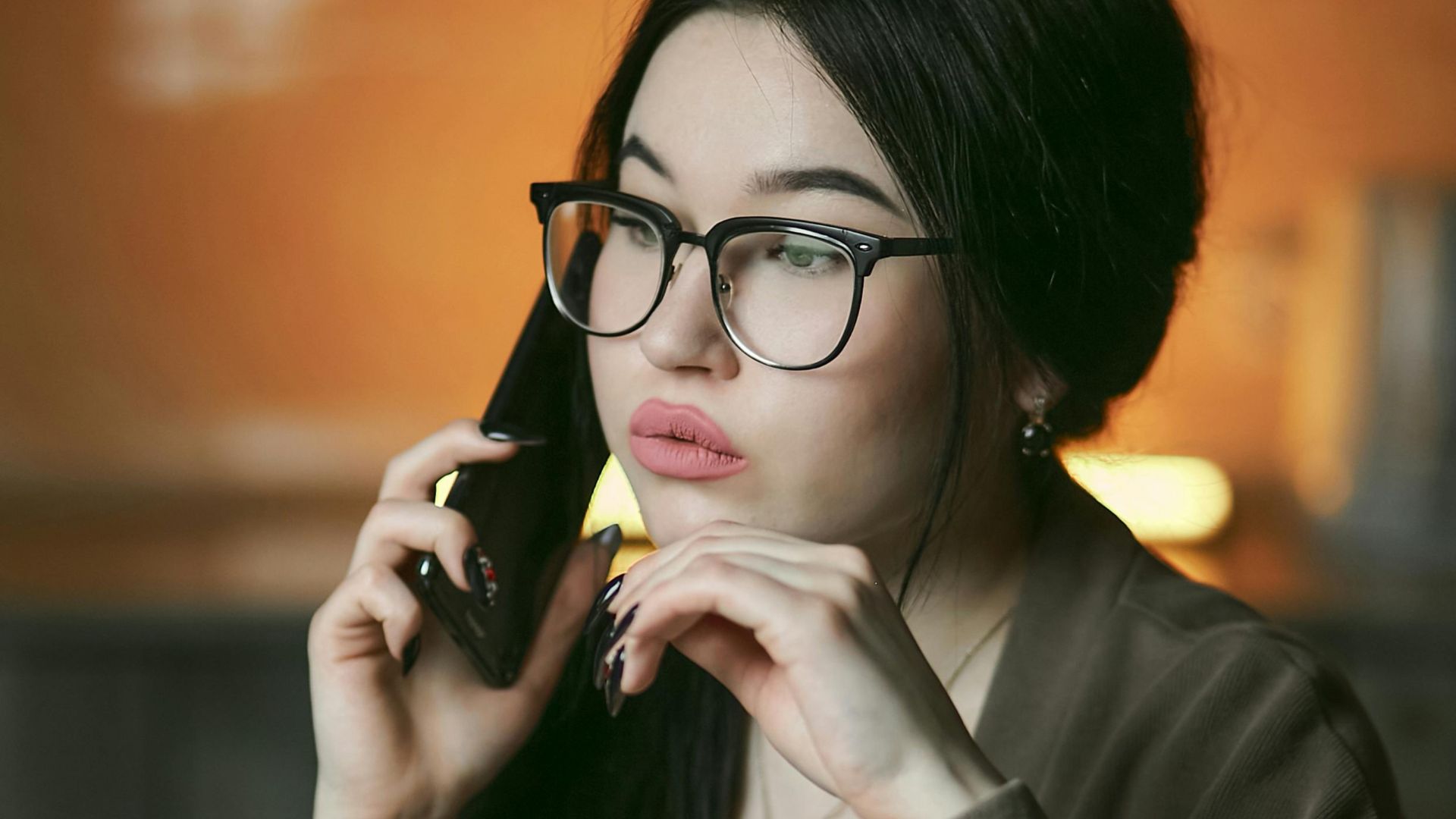 A woman with eyeglasses engaged in a phone call indoors, focused and thoughtful.