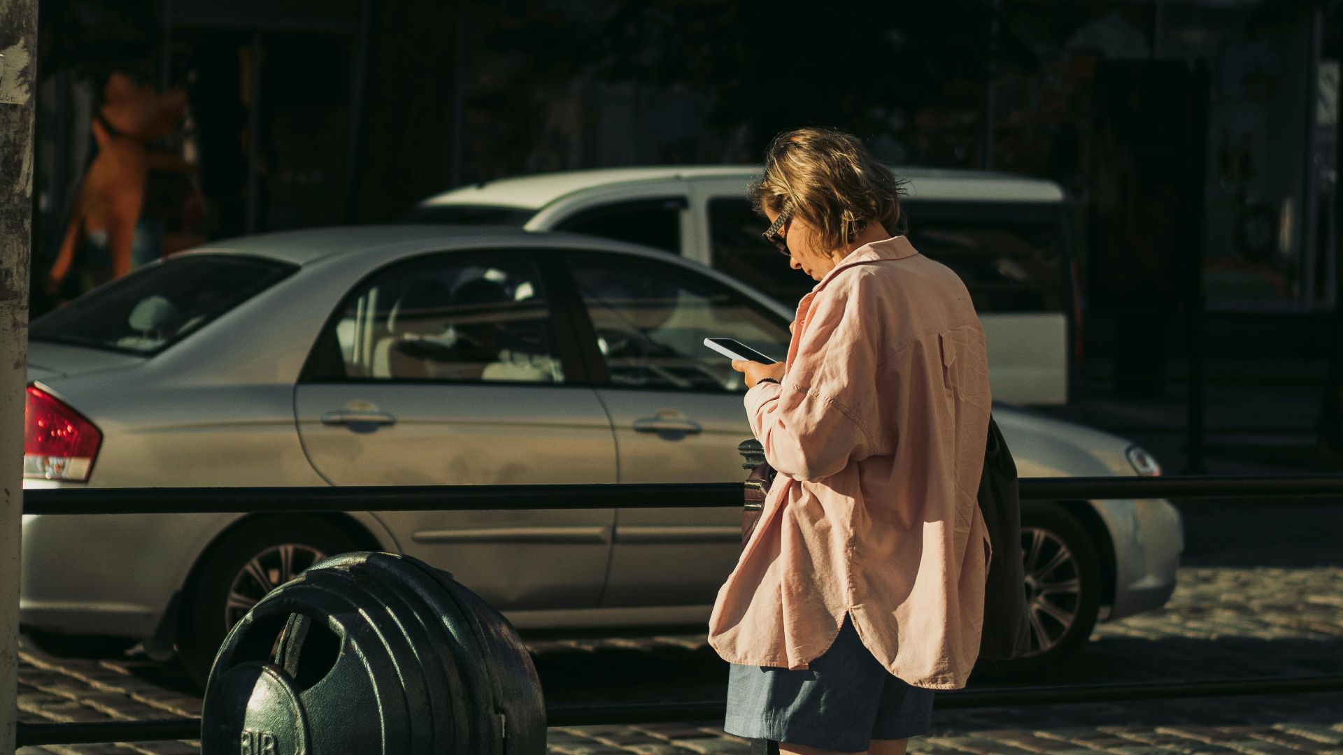 A woman standing on a sunlit street using her smartphone, surrounded by cars.