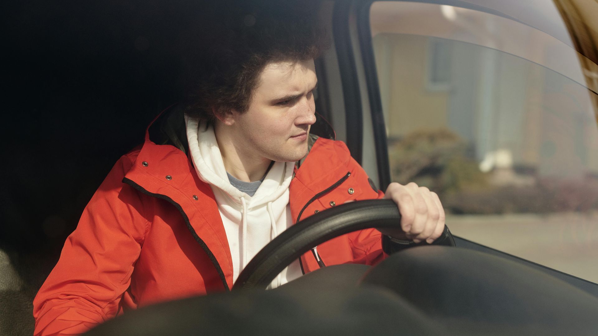 A young man in a red jacket steering a car with focus, enjoying a peaceful drive.