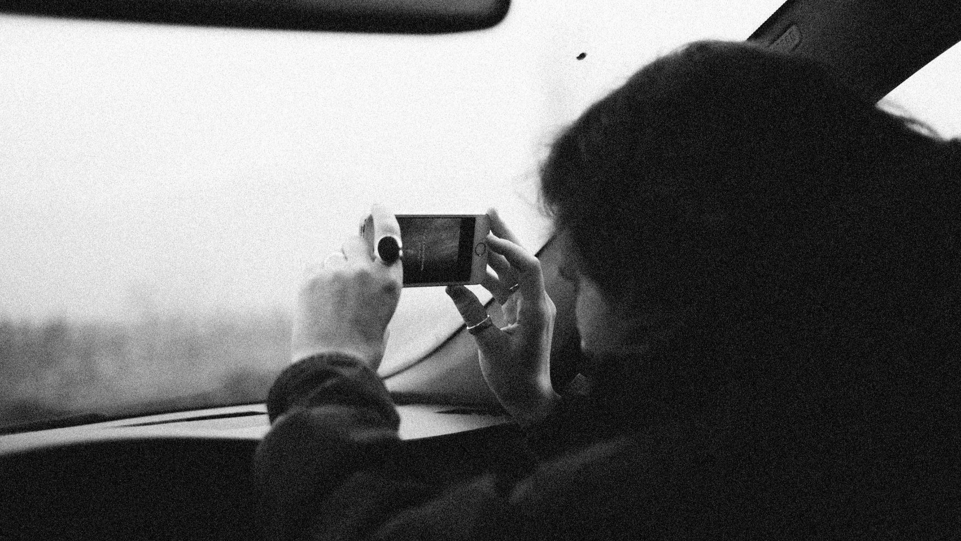 Monochrome image of a person capturing a view with a smartphone from a car.