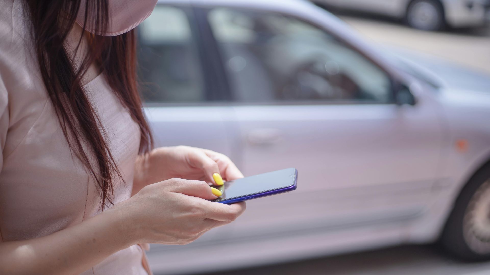 A close-up of a woman texting on a smartphone near a parked car outdoors.
