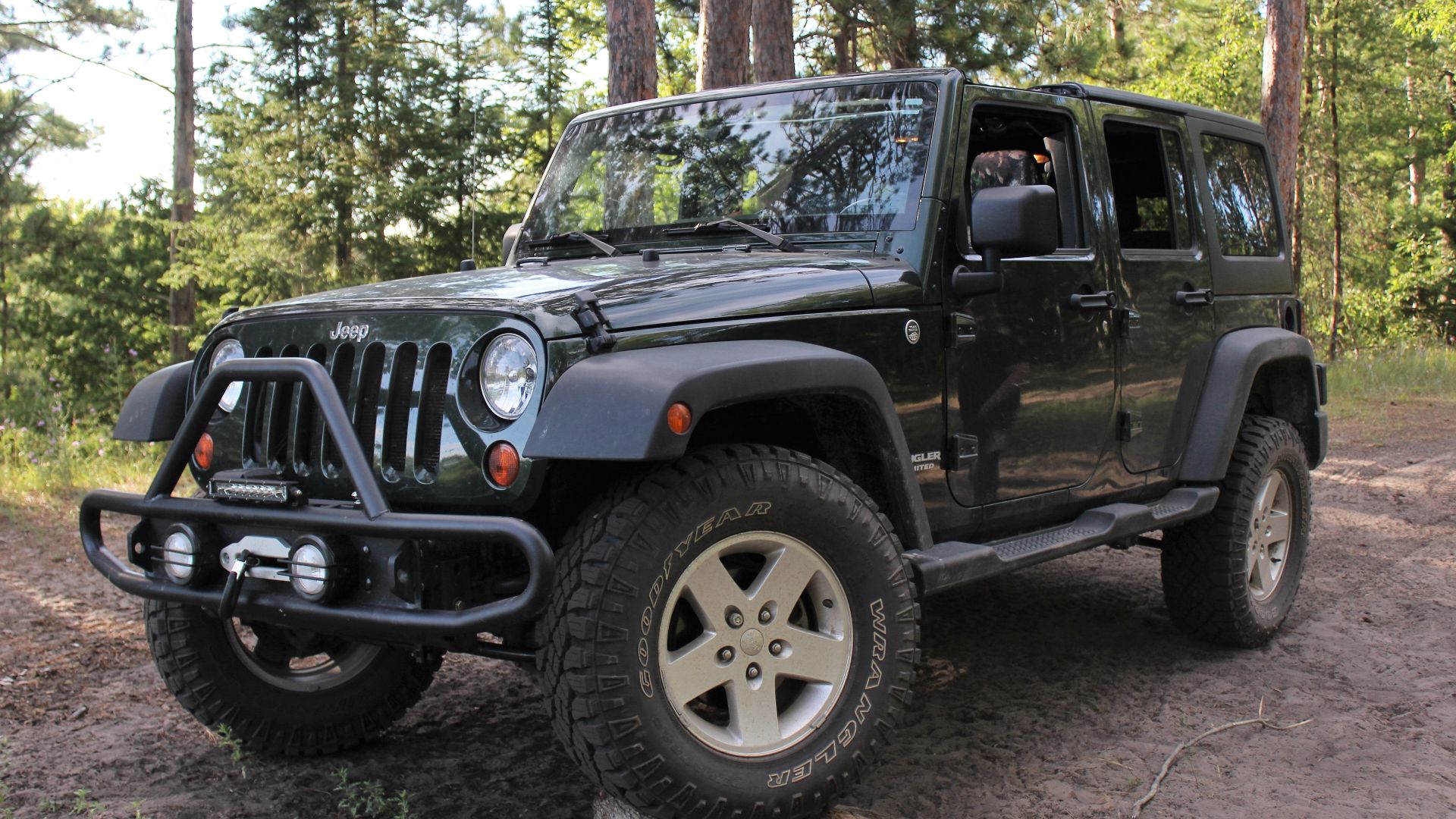 This is a picture of a dark green 2011 Jeep Wrangler. Its front left wheel is propped on some stacks of wood. The picture is taken in the forest near Black River on Fireline Road in Onaway, Michigan. The image corrections include: level adjusting, color correction, and minor cropping and rotation.