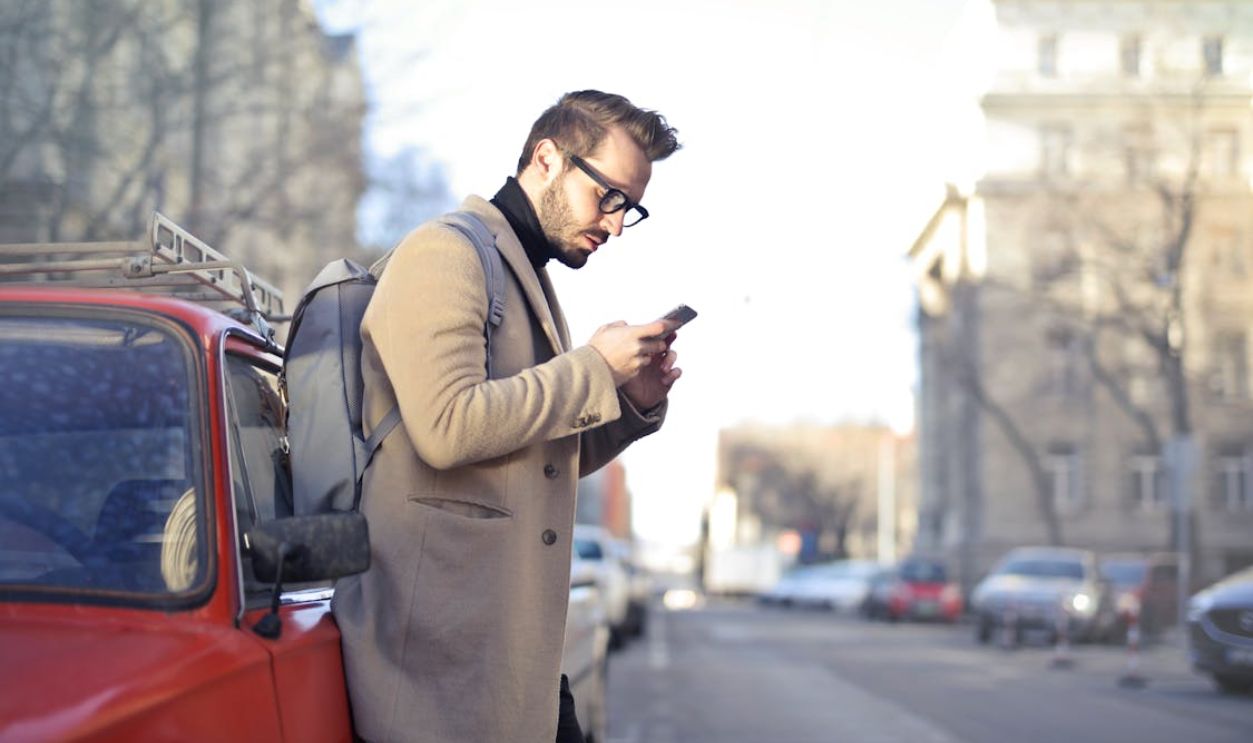 Man in Beige Coat Holding Phone Leaning on Red Vehicle