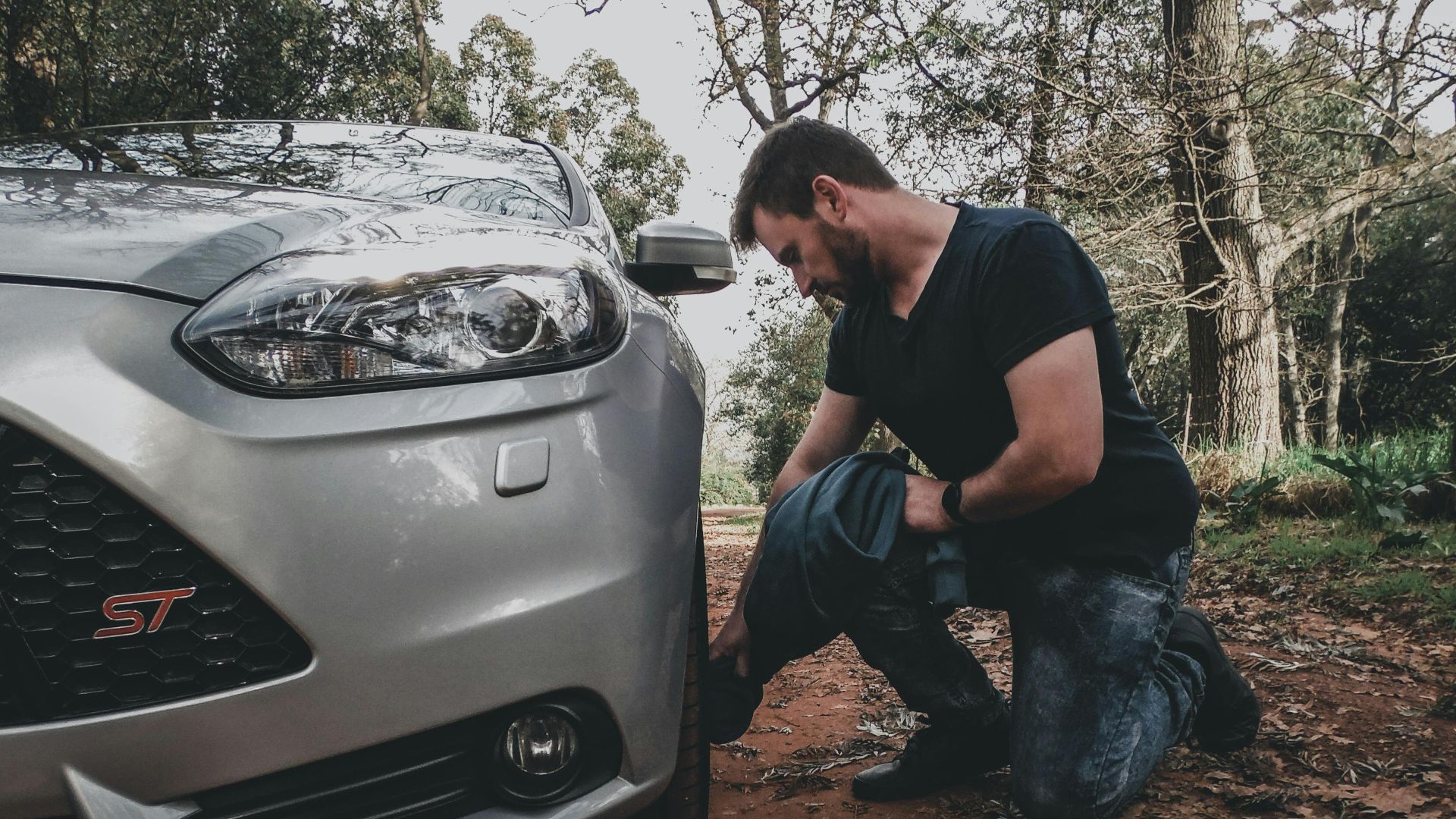 A man kneels to check the tire of a silver car in a forest, surrounded by trees.