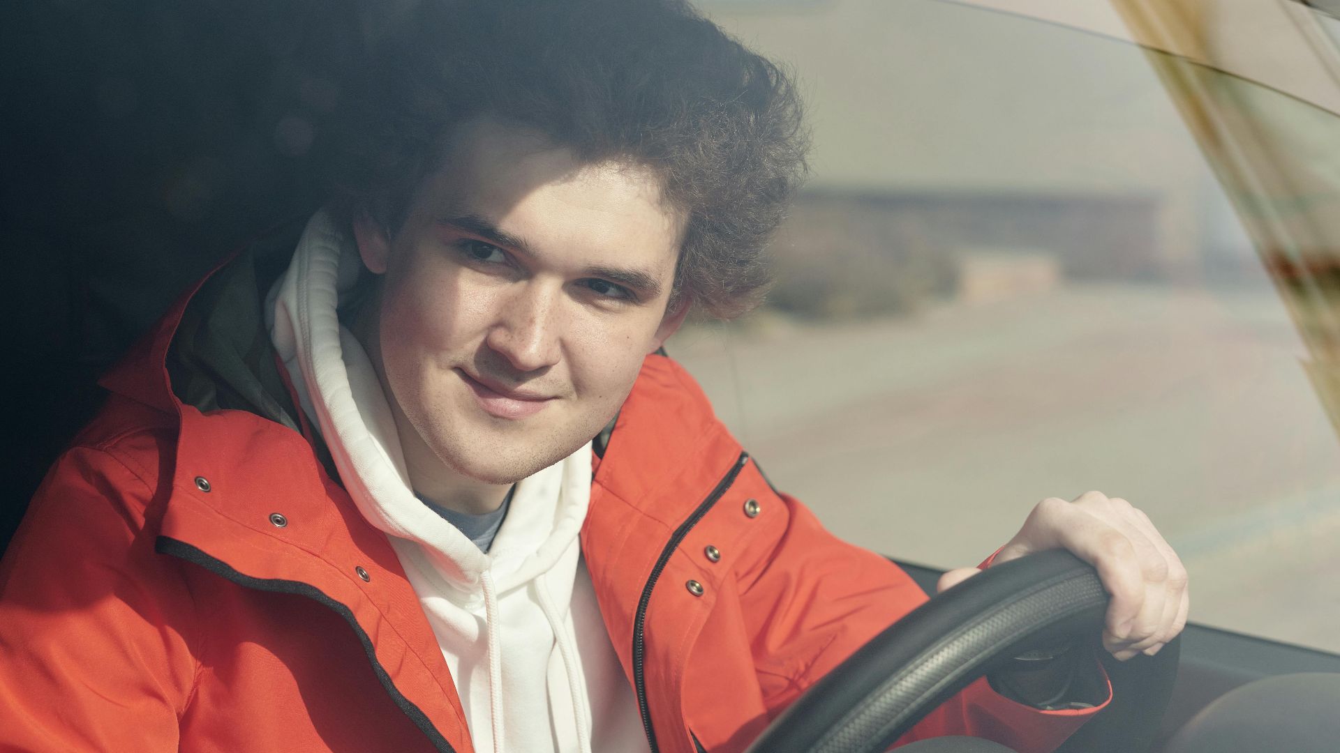 A young adult man smiling while sitting behind the steering wheel of a vehicle.