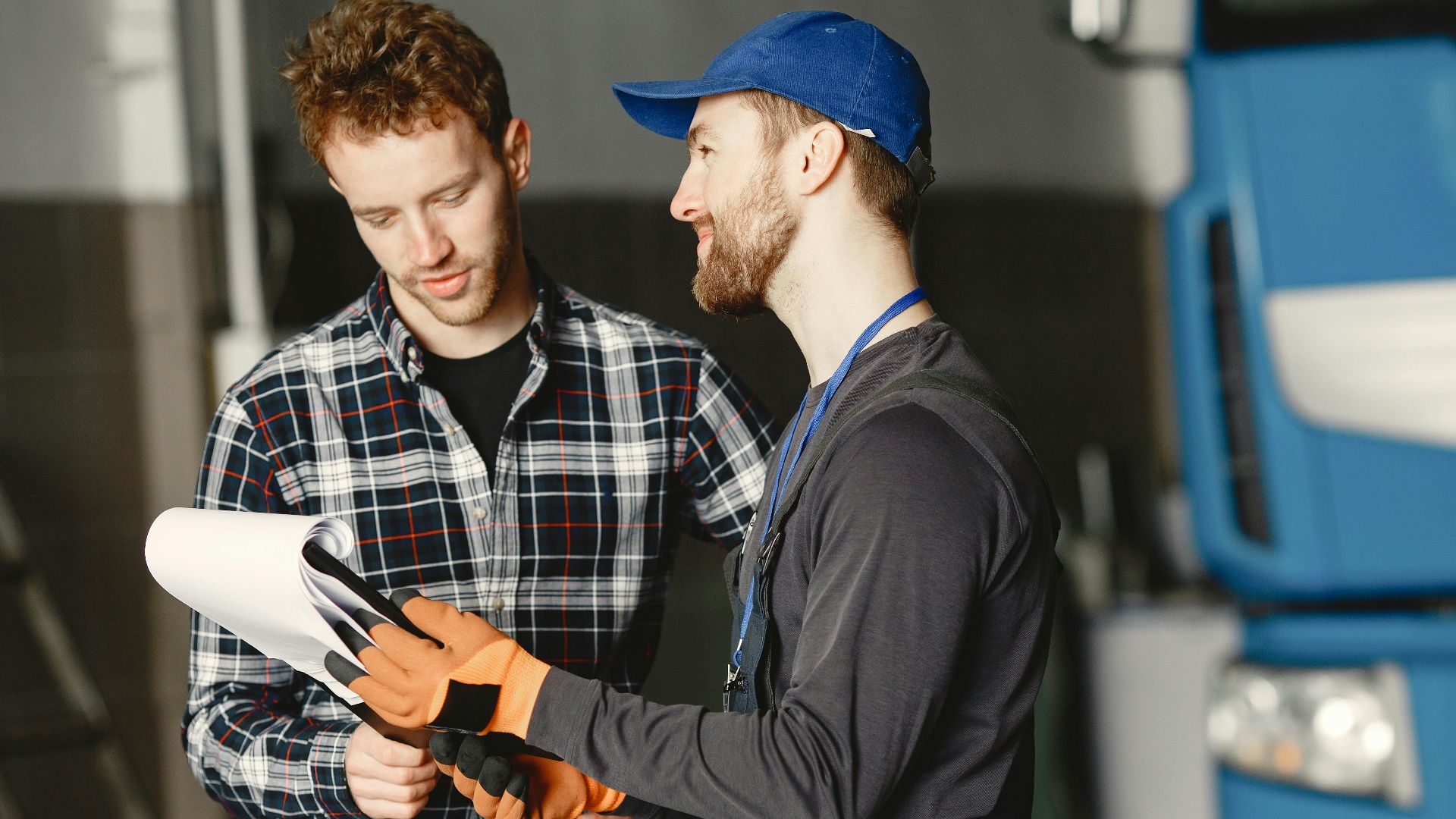 Mechanic in uniform consults with customer in garage setting beside a blue truck.