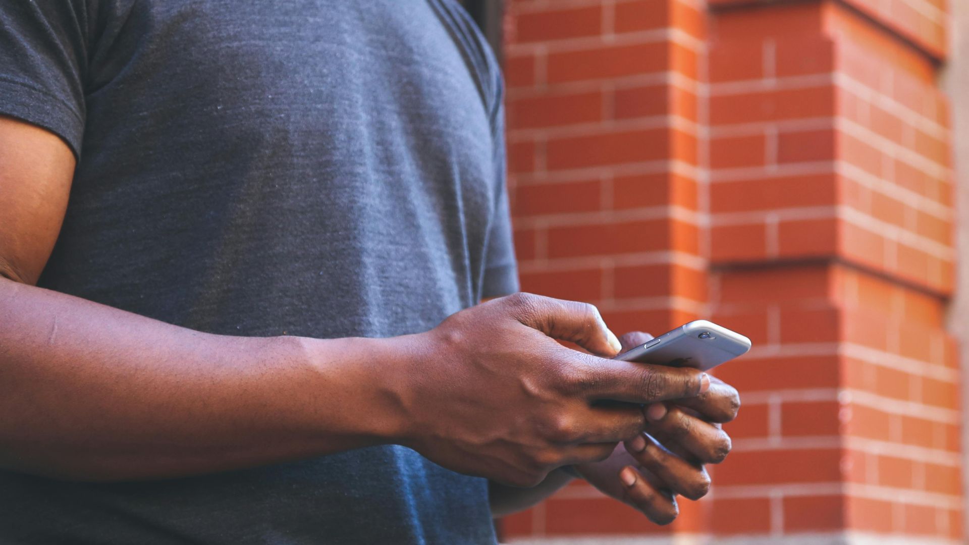 A smiling man checks his smartphone while leaning against a brick wall outdoors.