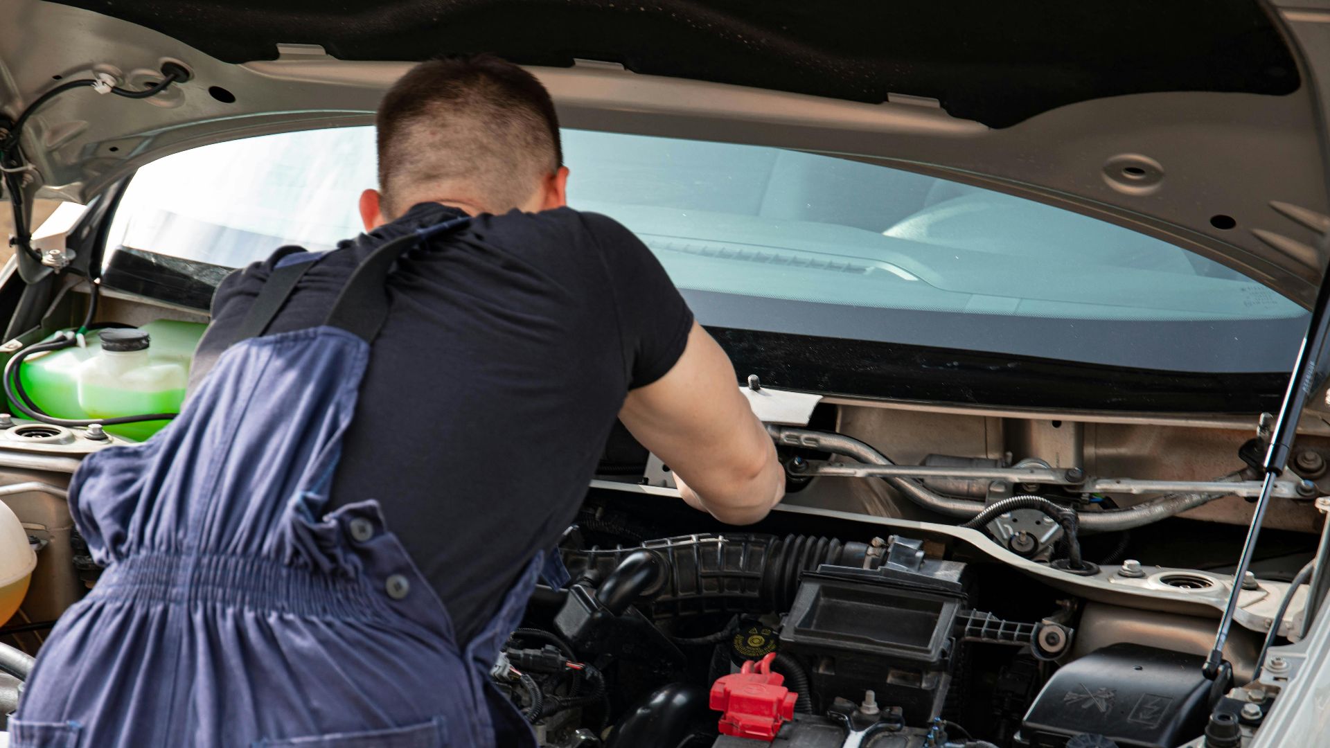 Mechanic in blue coveralls performing car engine maintenance outdoors.