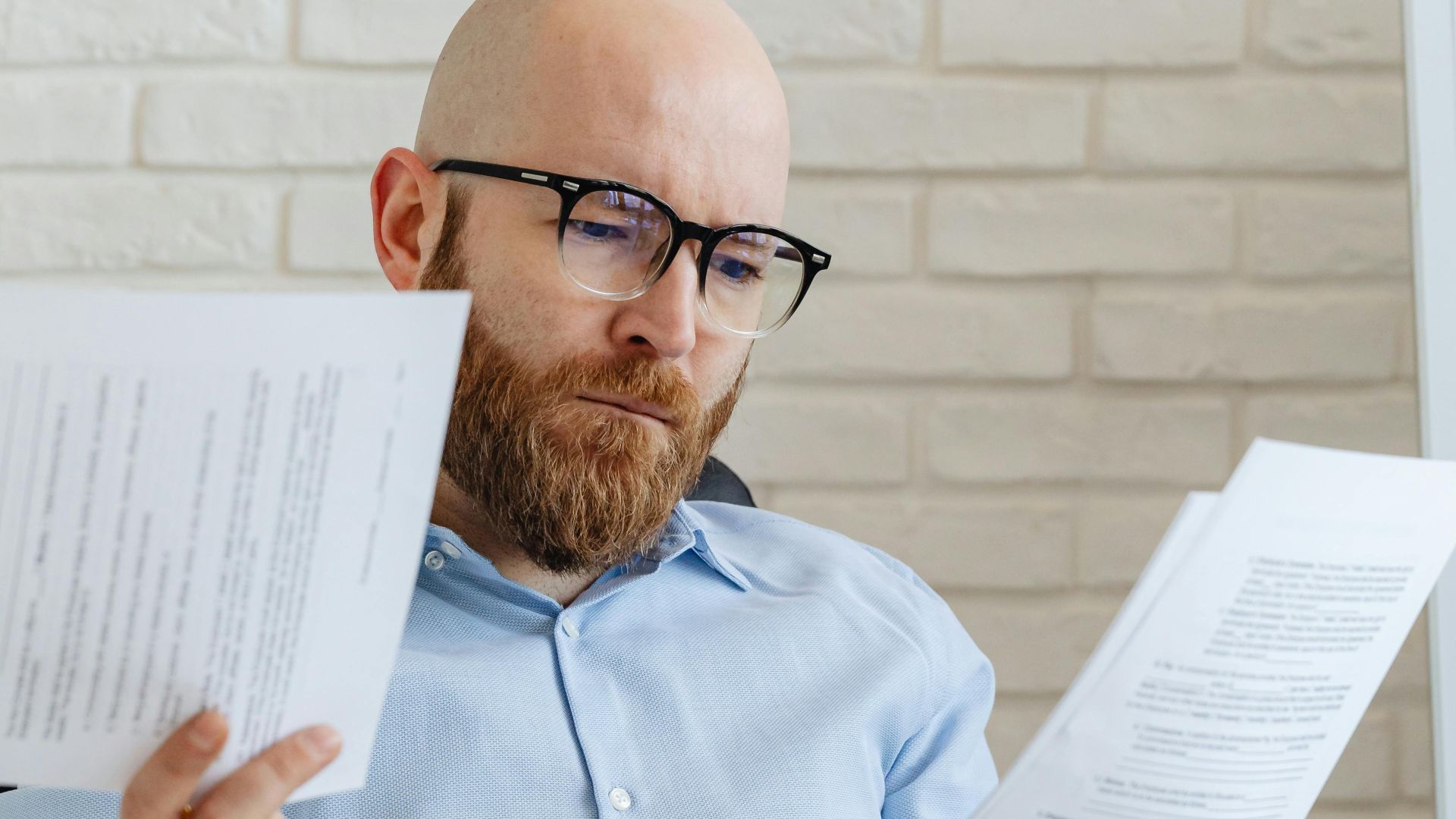 A serious-looking bald man with glasses reviews papers in an office against a brick wall.