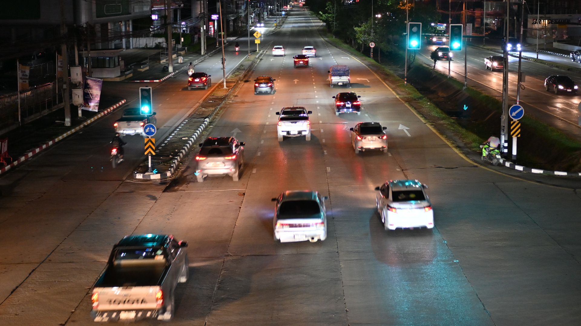 The photo shows a nighttime street scene in what appears to be a city or urban area. Several cars are visible driving on a wide road that's illuminated by street lights. There are traffic lights and signs visible at what seems to be an intersection. The surrounding buildings have some illuminated signs and advertisements, creating the typical colorful glow of a city at night. The road appears to be quite wide with multiple lanes, and various vehicles can be seen traveling in both directions. The overall scene captures the urban nightlife atmosphere with its artificial lighting against the dark sky.