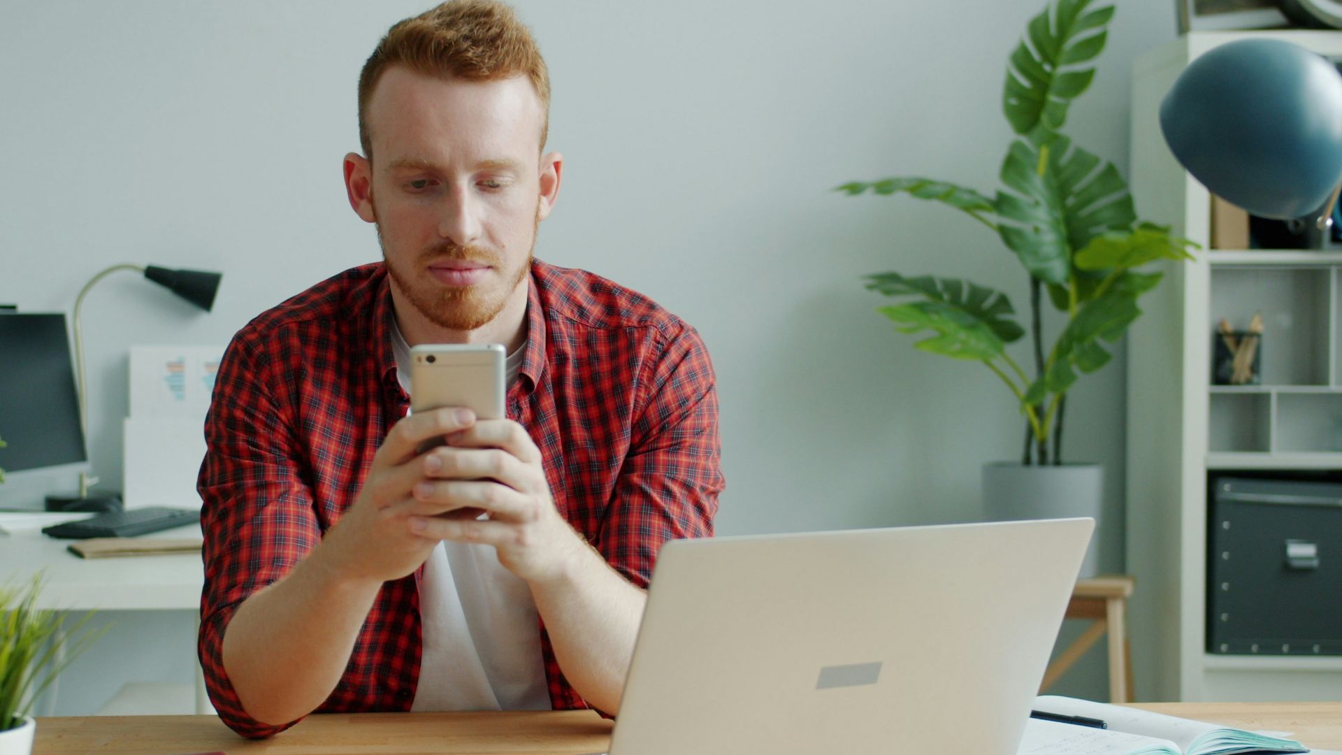 Man with red hair using smartphone at desk with laptop.