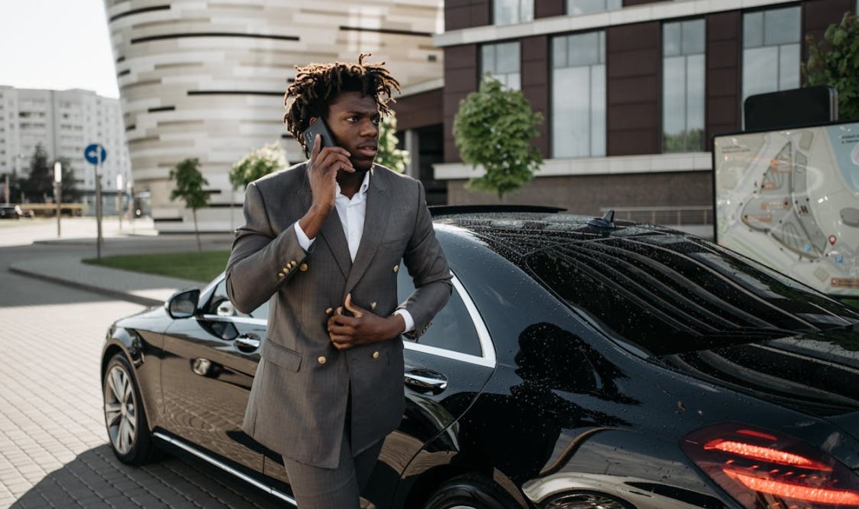 Confident businessman making a phone call while standing beside a luxury black car in an urban setting.