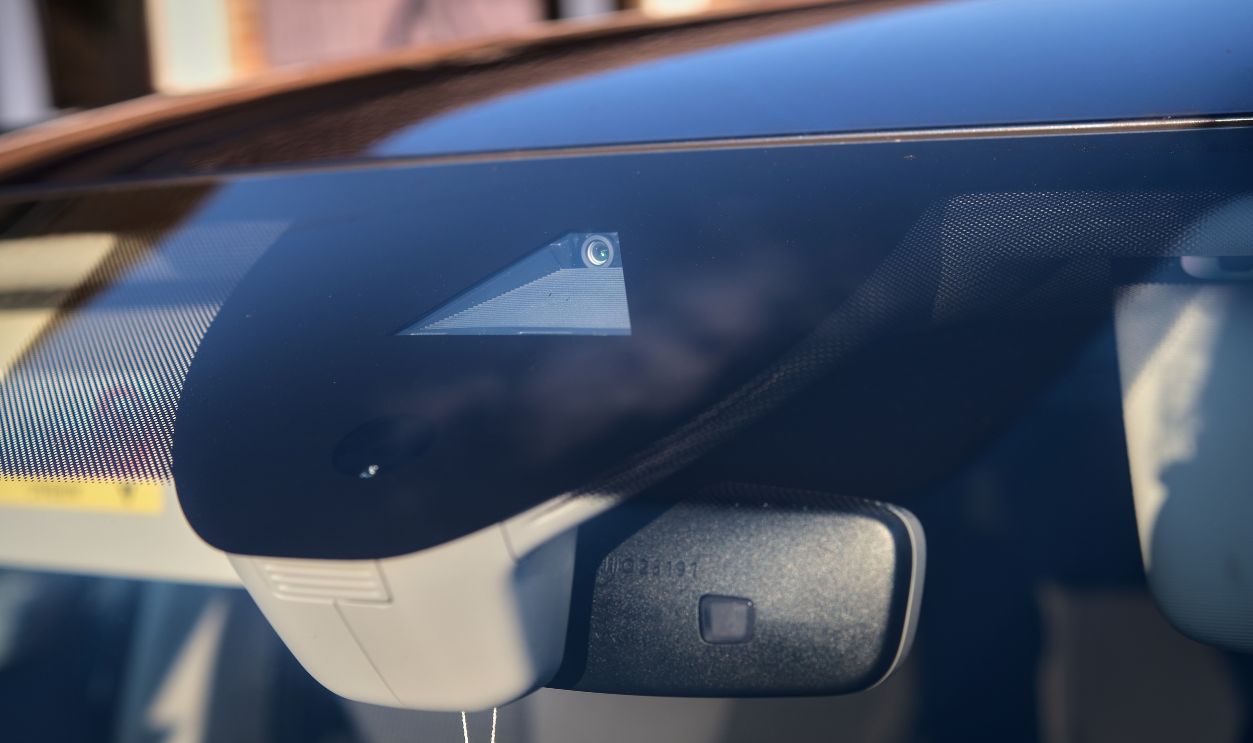 Close-up of a modern car's windshield with an integrated driver-assistance camera and sensor system, reflecting the surroundings in natural daylight.