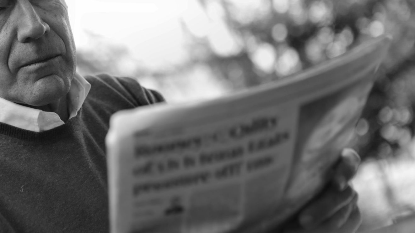 Elderly man reading a newspaper outdoors, wearing a blue sweater, showing serious expression.