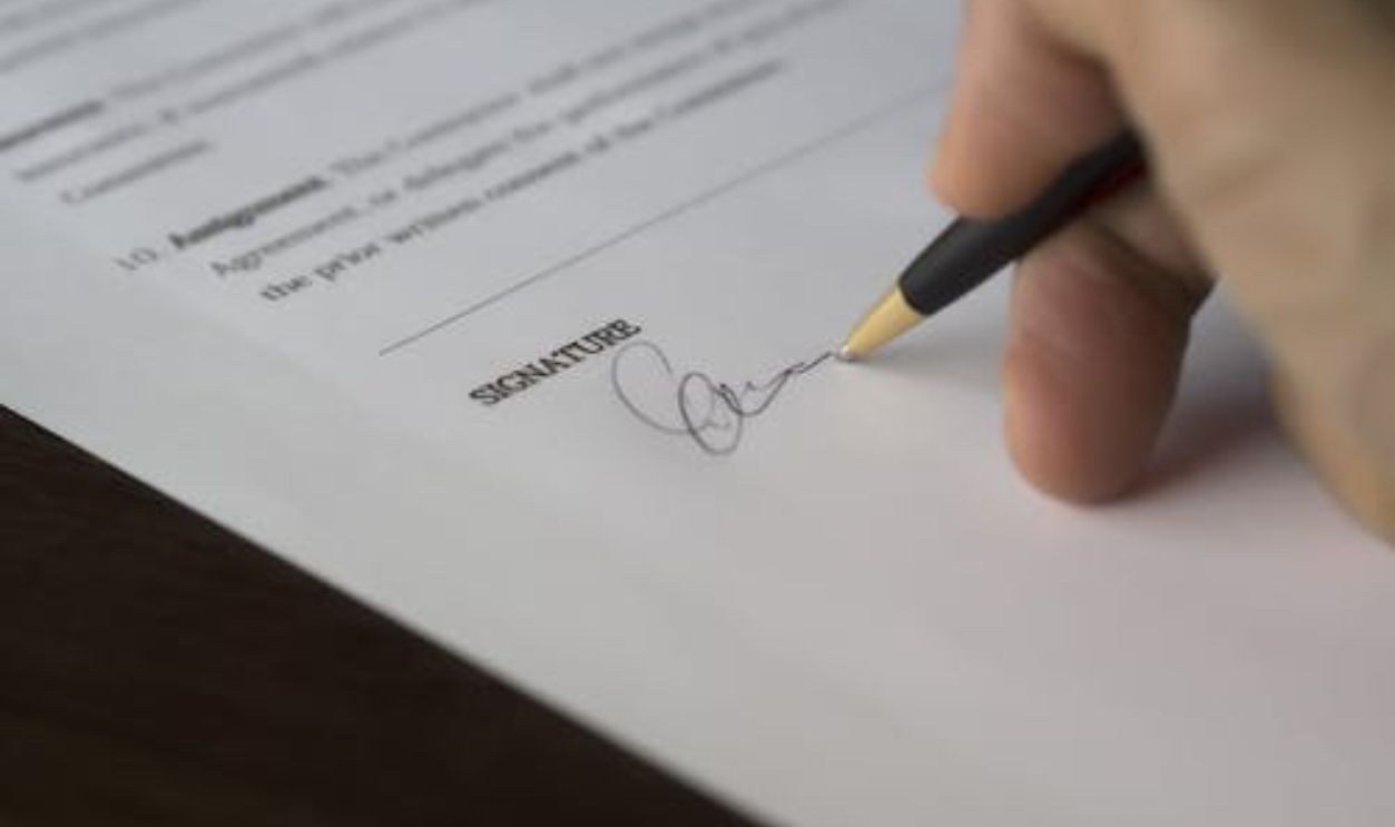 A hand signs a formal contract with a pen on a wooden desk