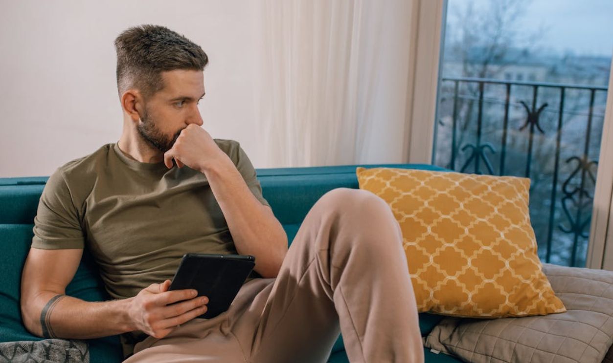 Bearded man sitting on a sofa, thoughtfully holding a tablet indoors