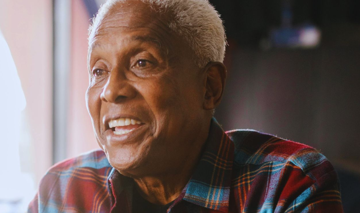 Elderly man expressing emotions while seated indoors at a wooden table with sunlight streaming in
