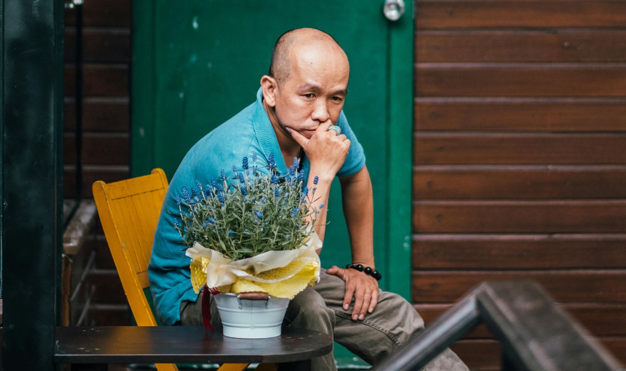 Bald man in casual shirt seated outdoors by a wooden cabin, contemplating