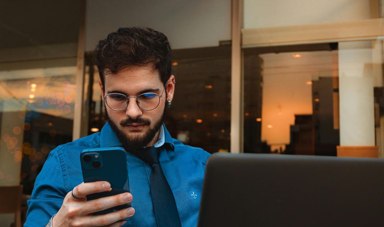 A man with a beard and eyeglasses focuses on his smartphone, sitting indoors with a laptop