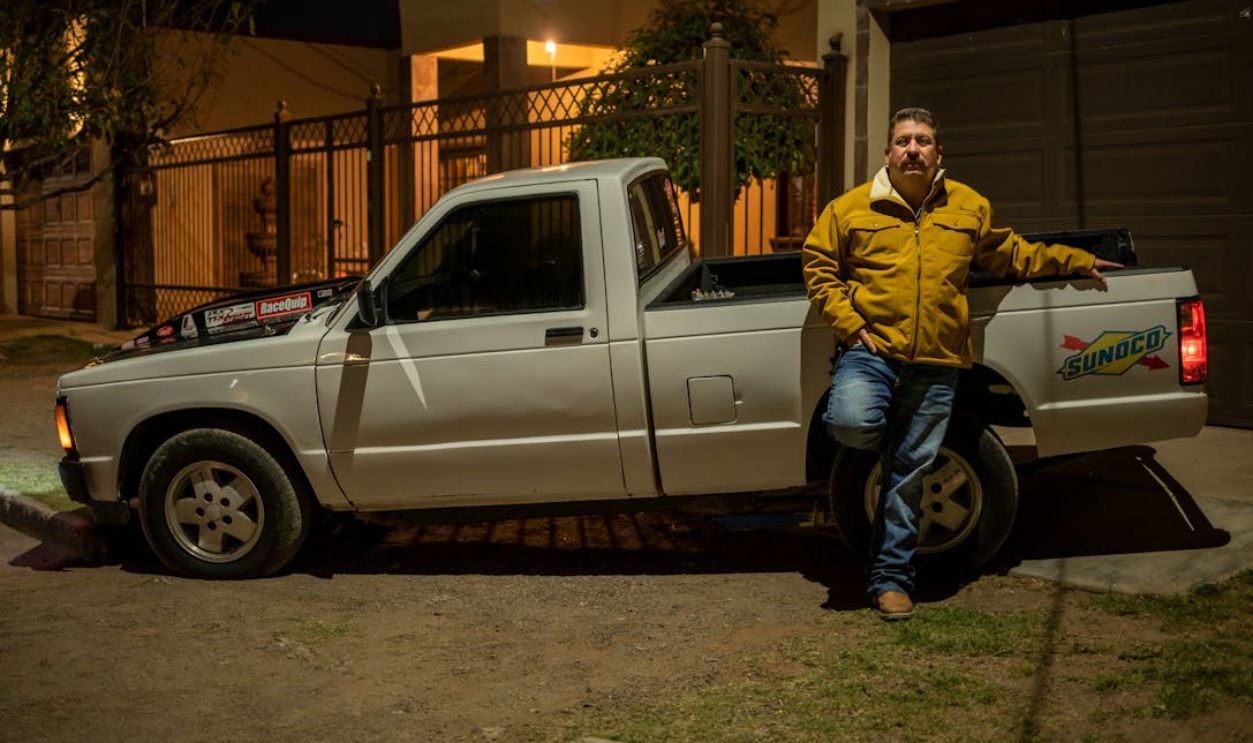 A man in a yellow jacket leaning on a white pickup truck at night in an urban area.