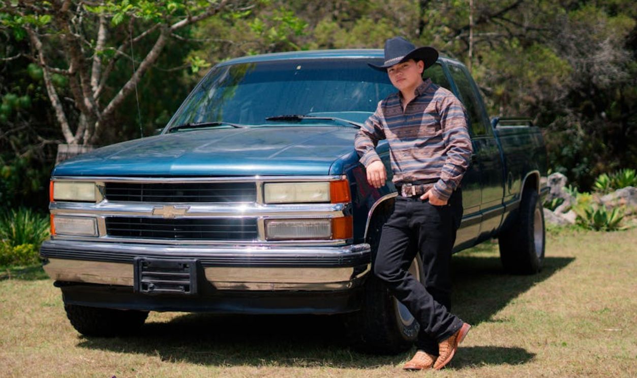 A cowboy in a hat relaxes against a classic blue truck outdoors in a sunny field