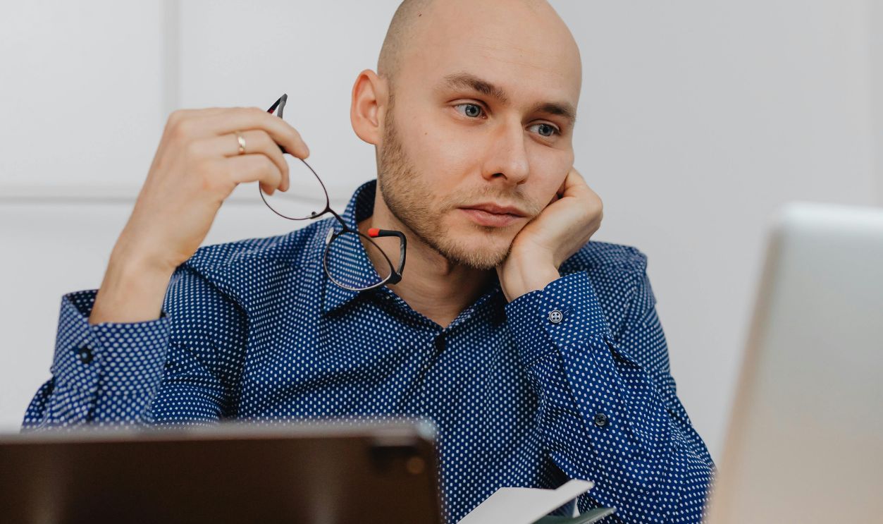 Thoughtful businessman holding glasses while working at his office desk using a laptop
