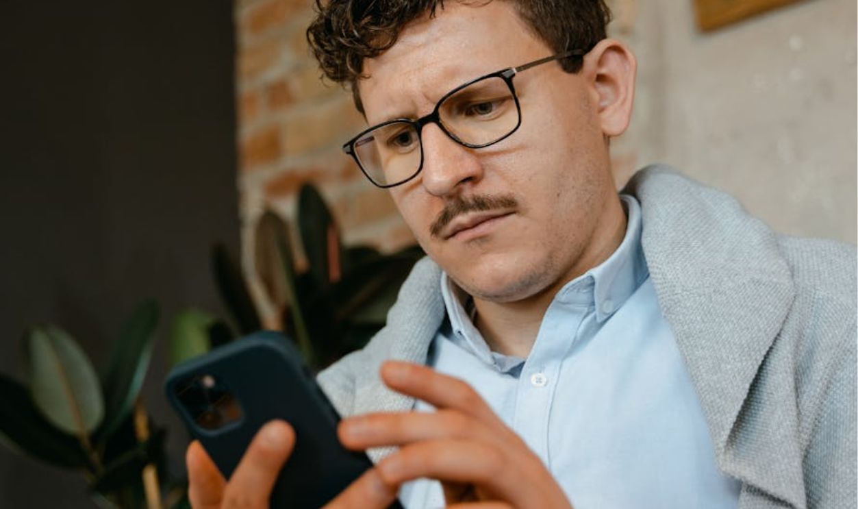 A focused man with glasses using a smartphone indoors next to a brick wall