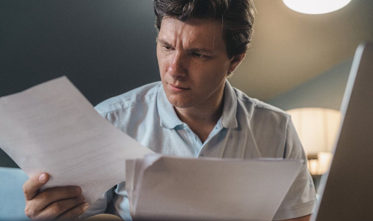 A man intently examines papers, seated indoors under warm lighting, focusing on his work