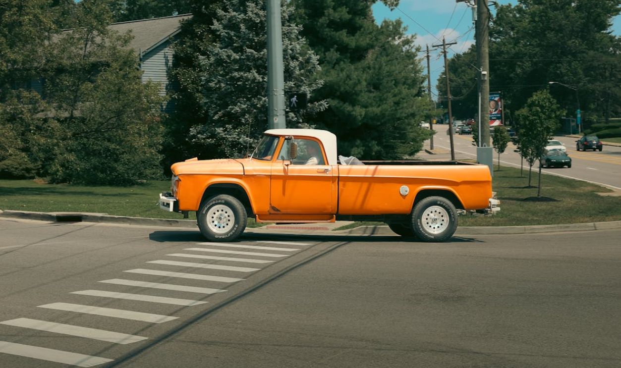 Classic orange pickup truck driving through a suburban intersection on a sunny day.