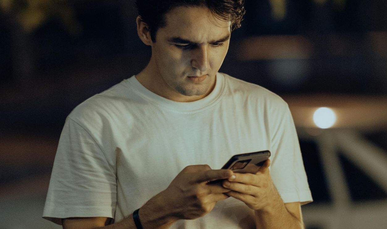 A young man in a white shirt stands outdoors using his smartphone during nighttime