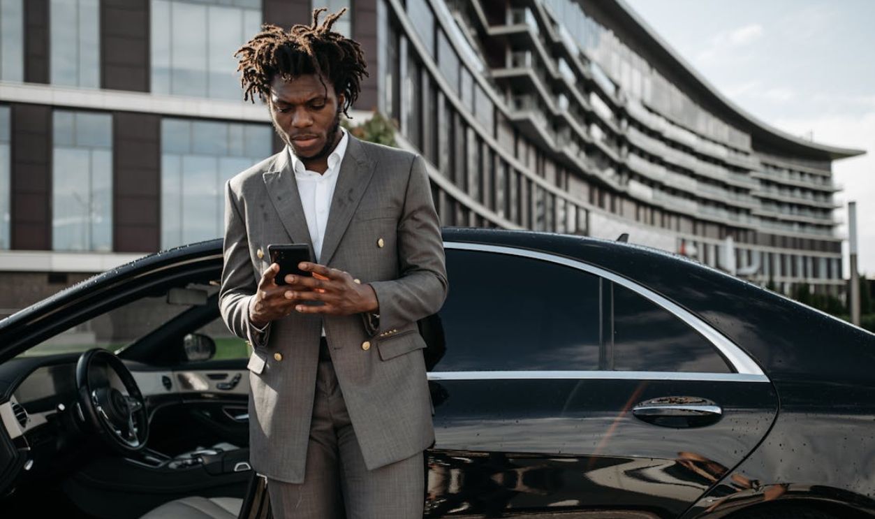 Professional man in a gray suit using his phone next to a luxury car.