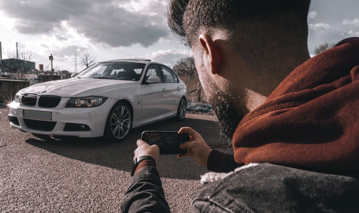 A man takes a picture of a white luxury car using a smartphone outdoors under a cloudy sky.