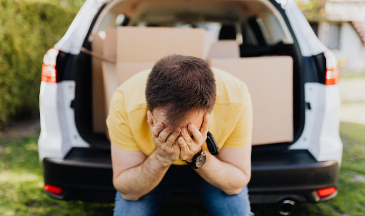 A distressed man sitting by a car trunk filled with cardboard boxes, expressing stress.