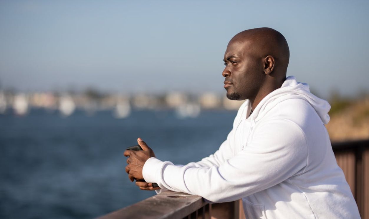 A man leans on a railing, overlooking a sunny waterfront while wearing a white hoodie.