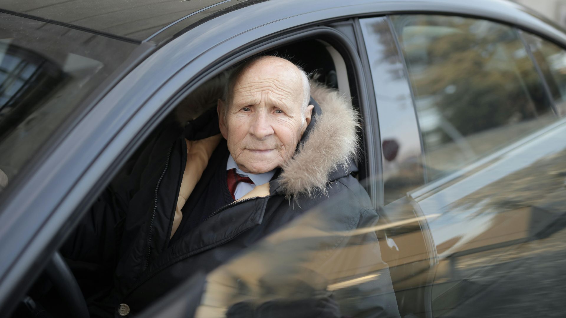 Senior man driving a car, wearing a winter coat, captured through the vehicle window.