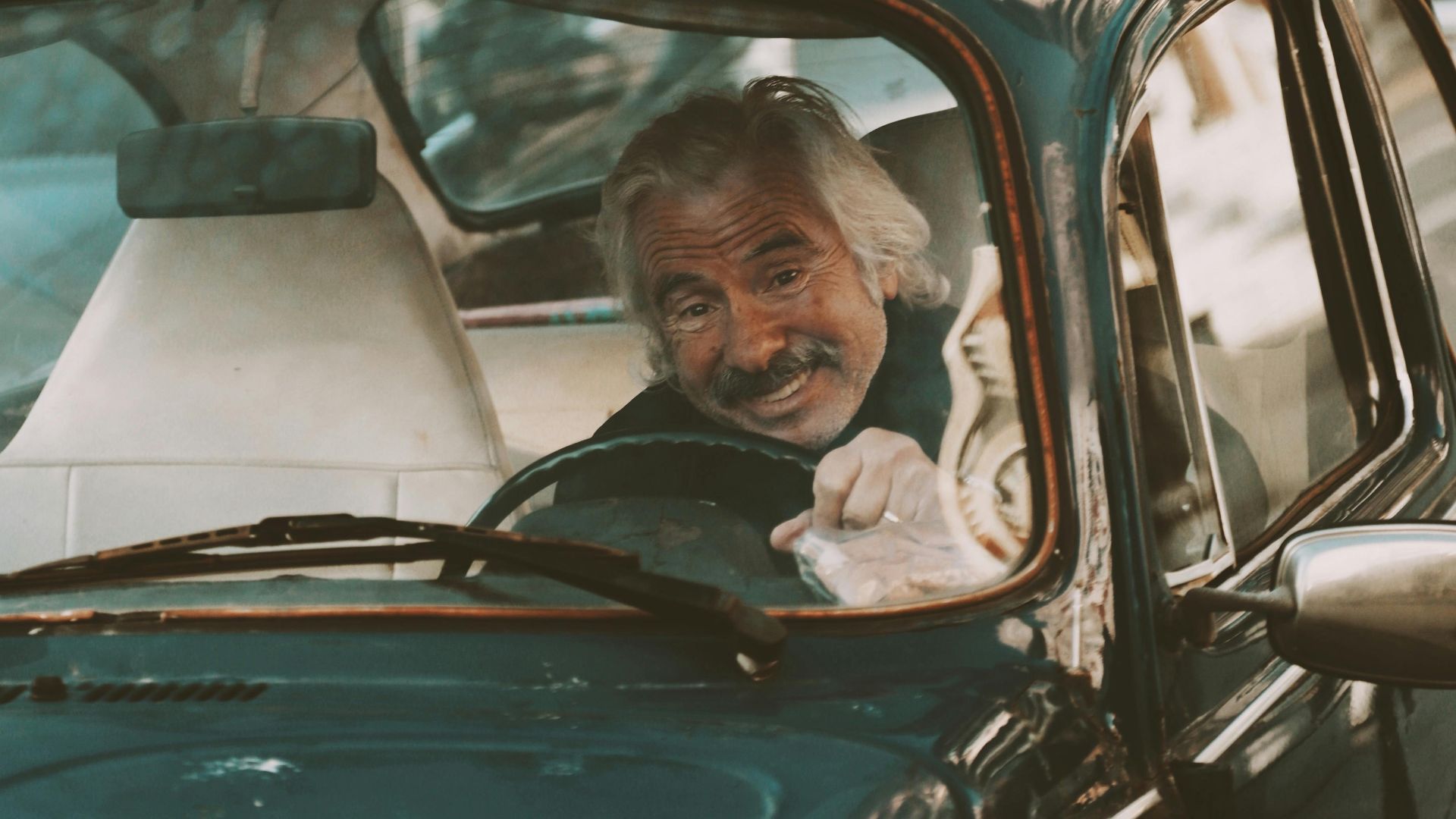 Elderly man smiling while driving a classic Volkswagen Beetle in traffic.