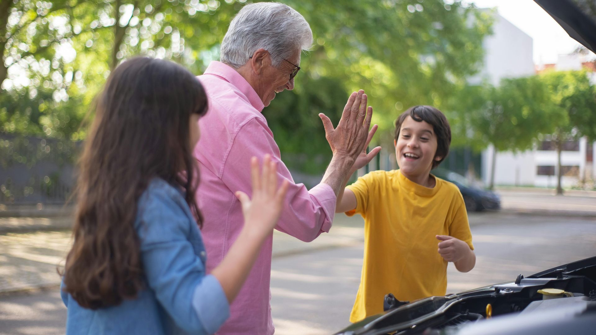 Cheerful grandfather and grandchildren high-five near car outdoors, enjoying a sunny day together.