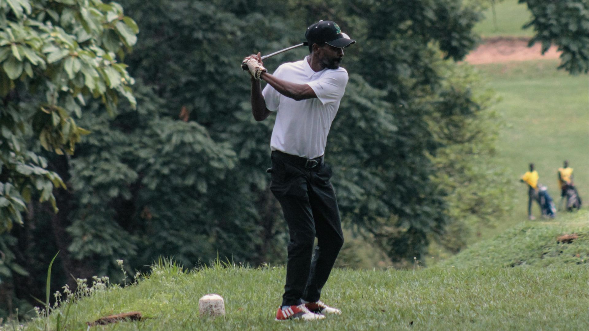 A golfer in action swinging his club on a green golf course surrounded by trees.