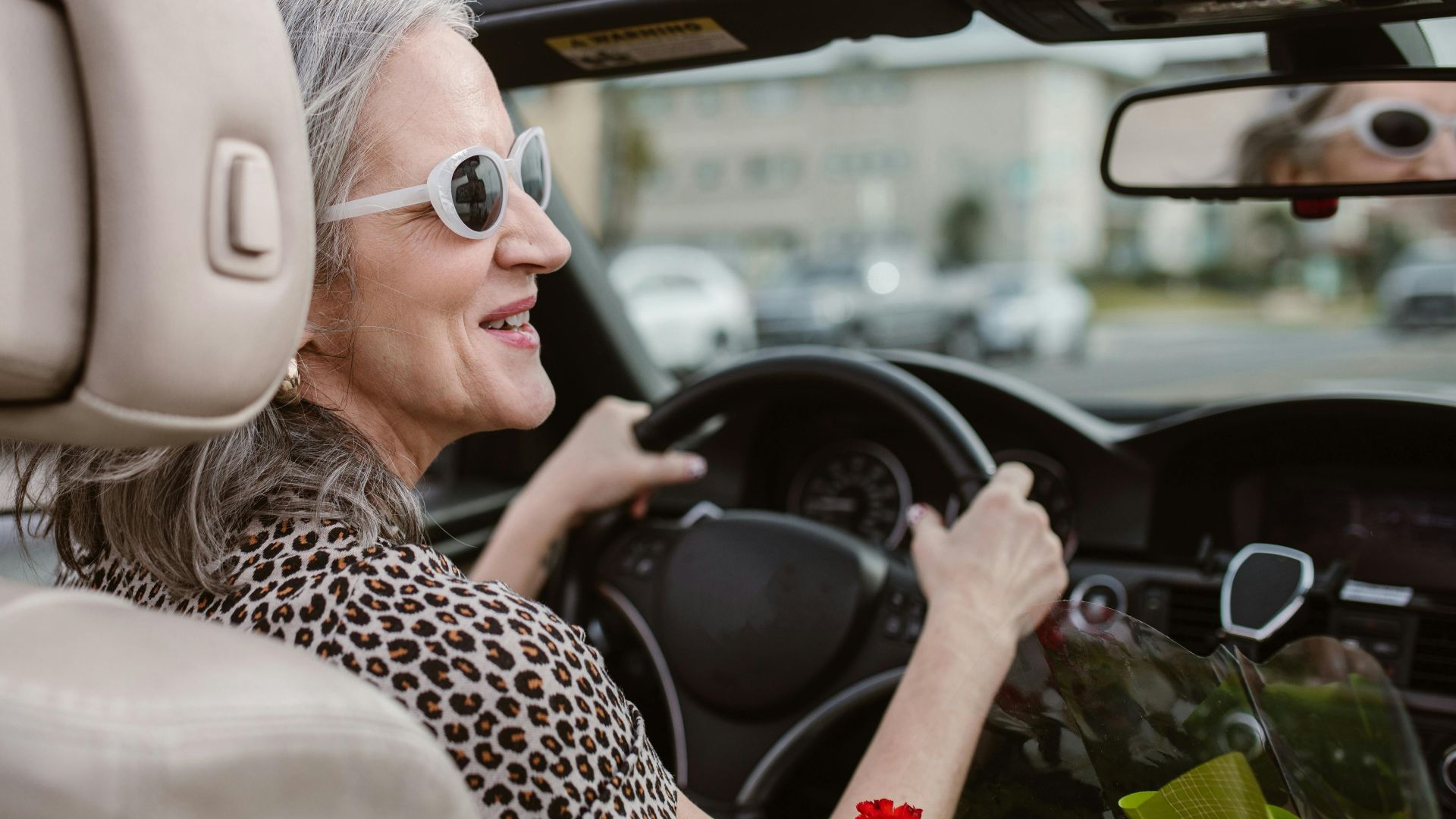 Senior woman driving a convertible, smiling and wearing sunglasses on a sunny day.