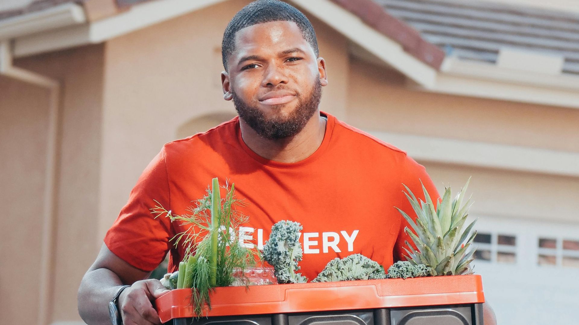 Delivery man carrying a box of groceries with pineapples outside a house.