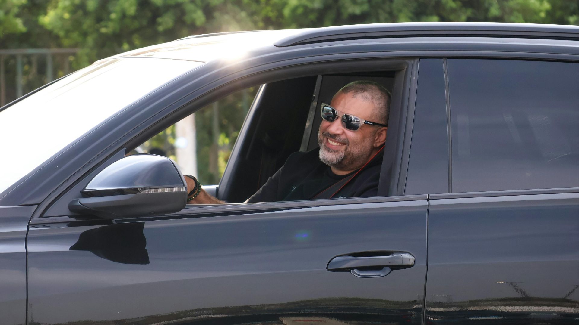 A smiling man with sunglasses driving a black car outdoors on a sunny day.