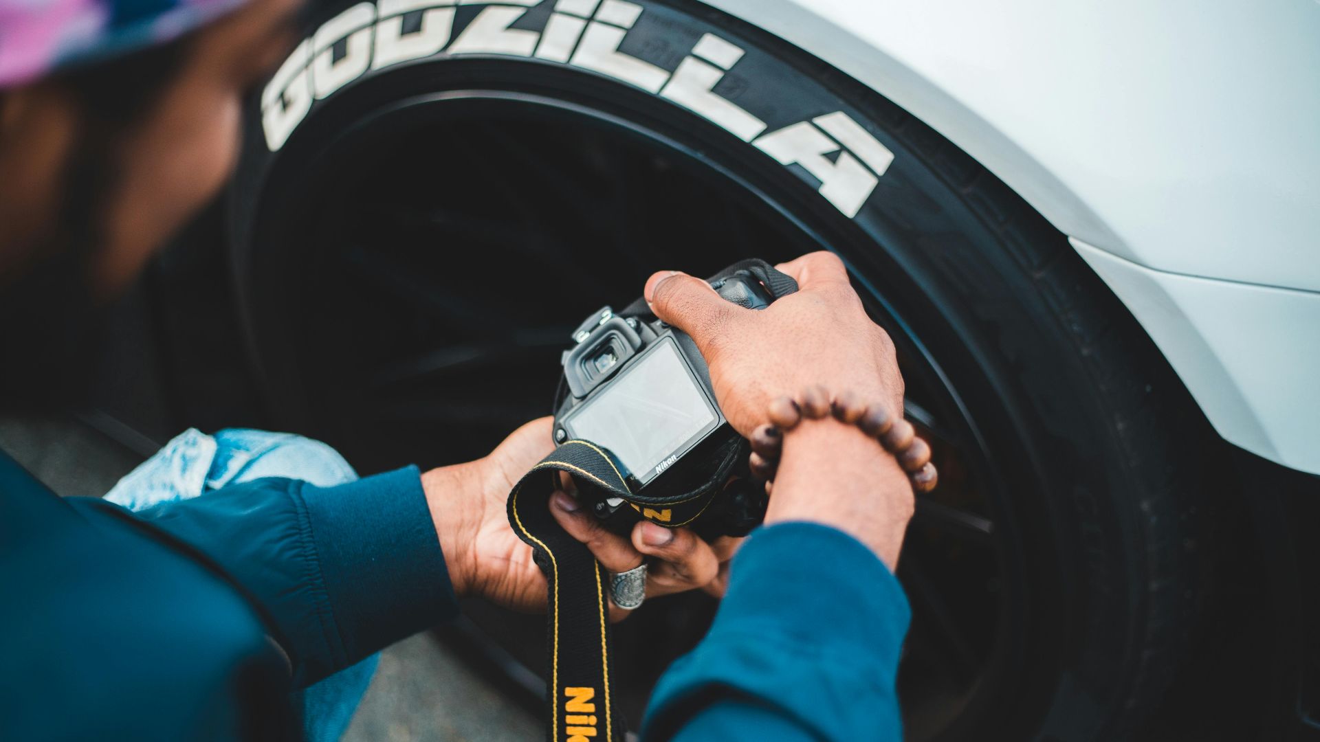 From above back view of anonymous bearded photographer in casual wear taking photo of contemporary auto wheel on digital camera with display and strap on pavement of city street