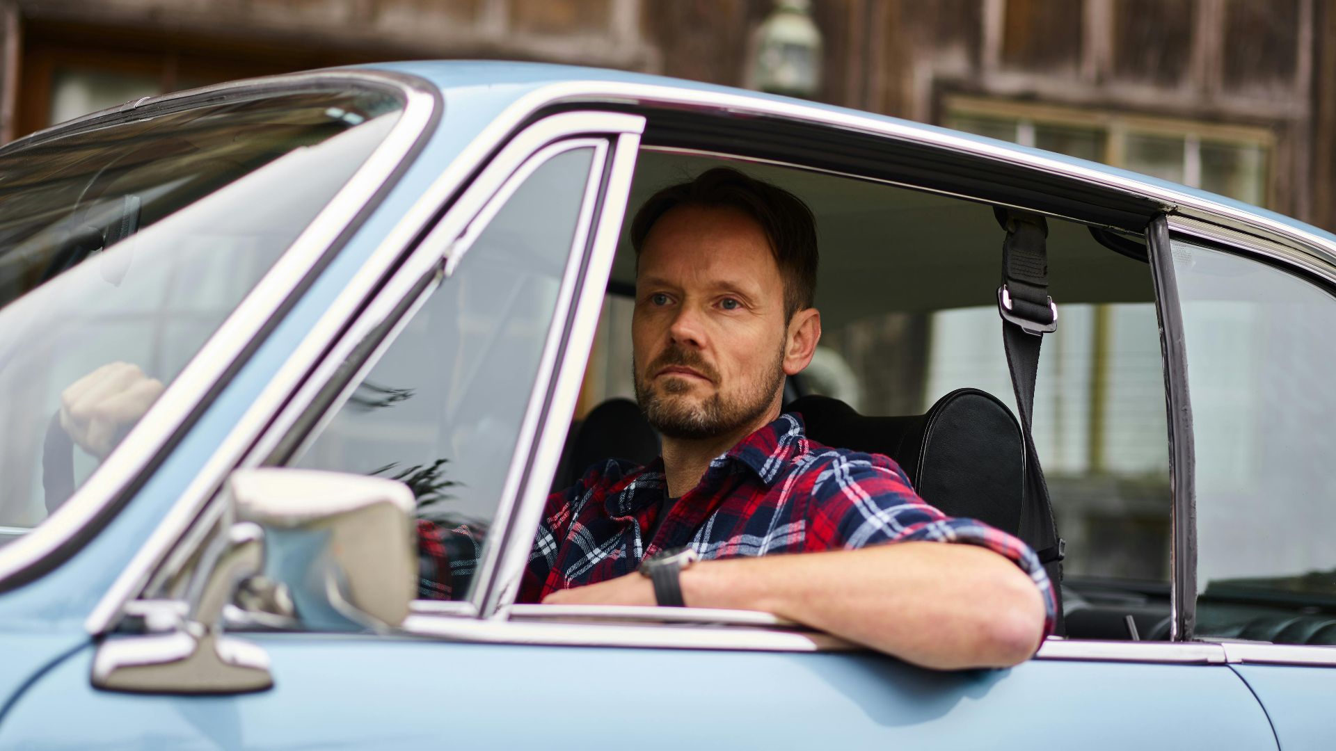 Adult thoughtful bearded driver in wristwatch and casual clothes driving blue automobile while passing by old building and looking away in daylight