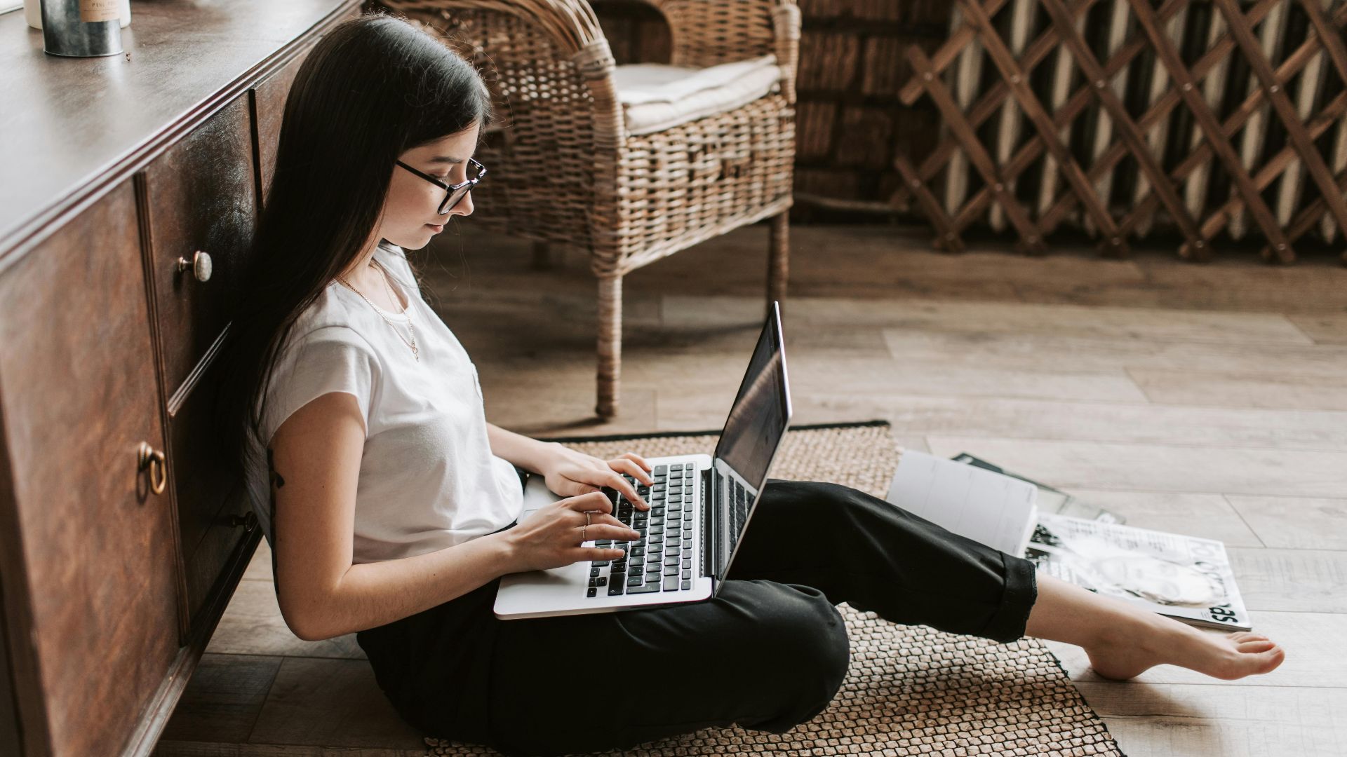 Side view of happy young woman with long dark hair in eyeglasses typing on keyboard of computer while sitting on floor in modern living room
