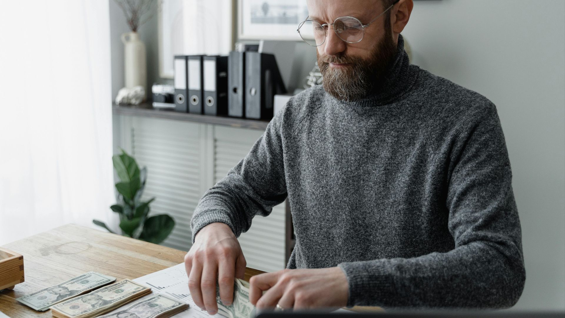 An adult man in a gray sweater counting dollar bills at a wooden desk, showcasing financial management.
