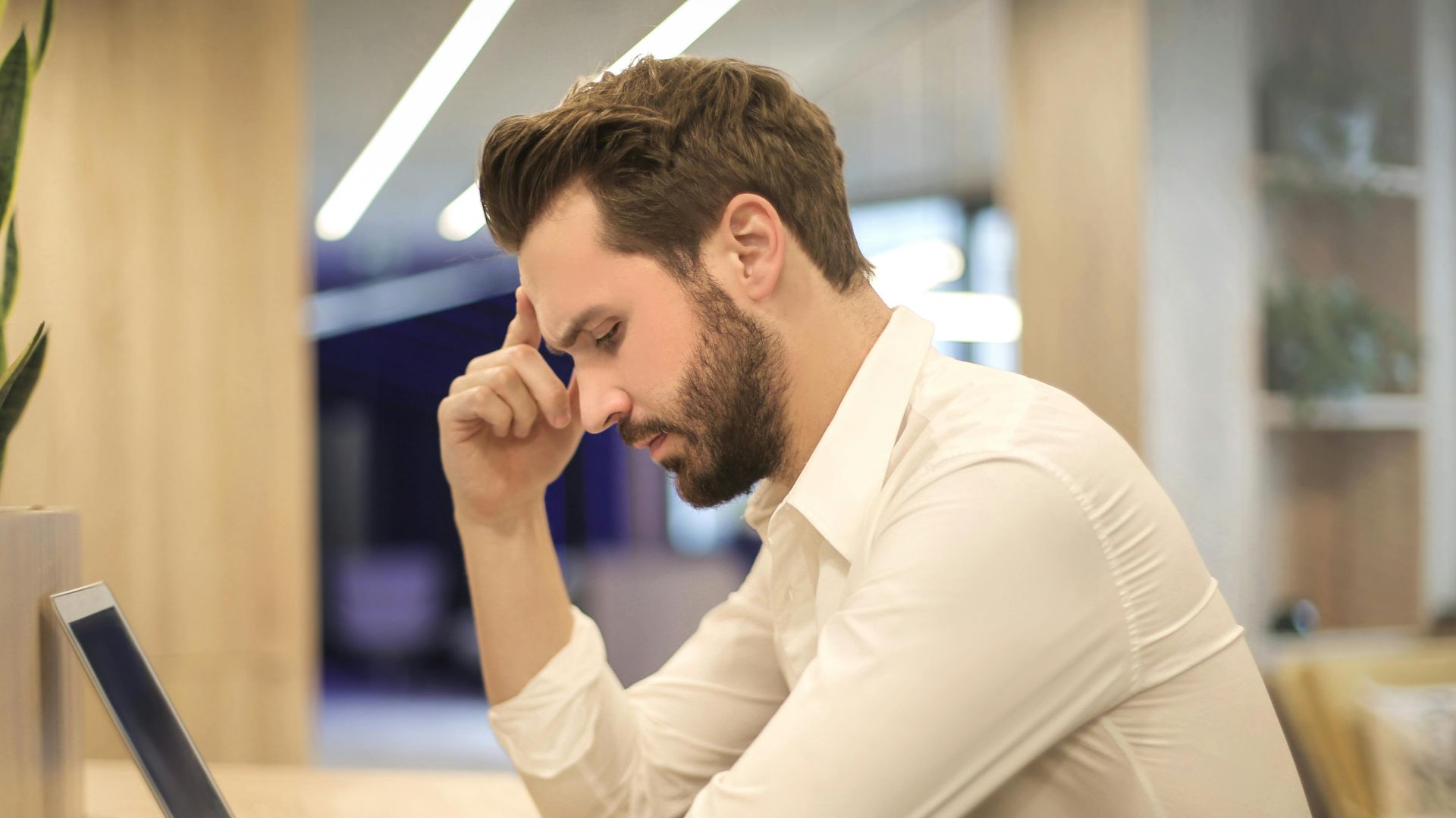 A thoughtful man in a shirt works on his laptop at a modern indoor office space.
