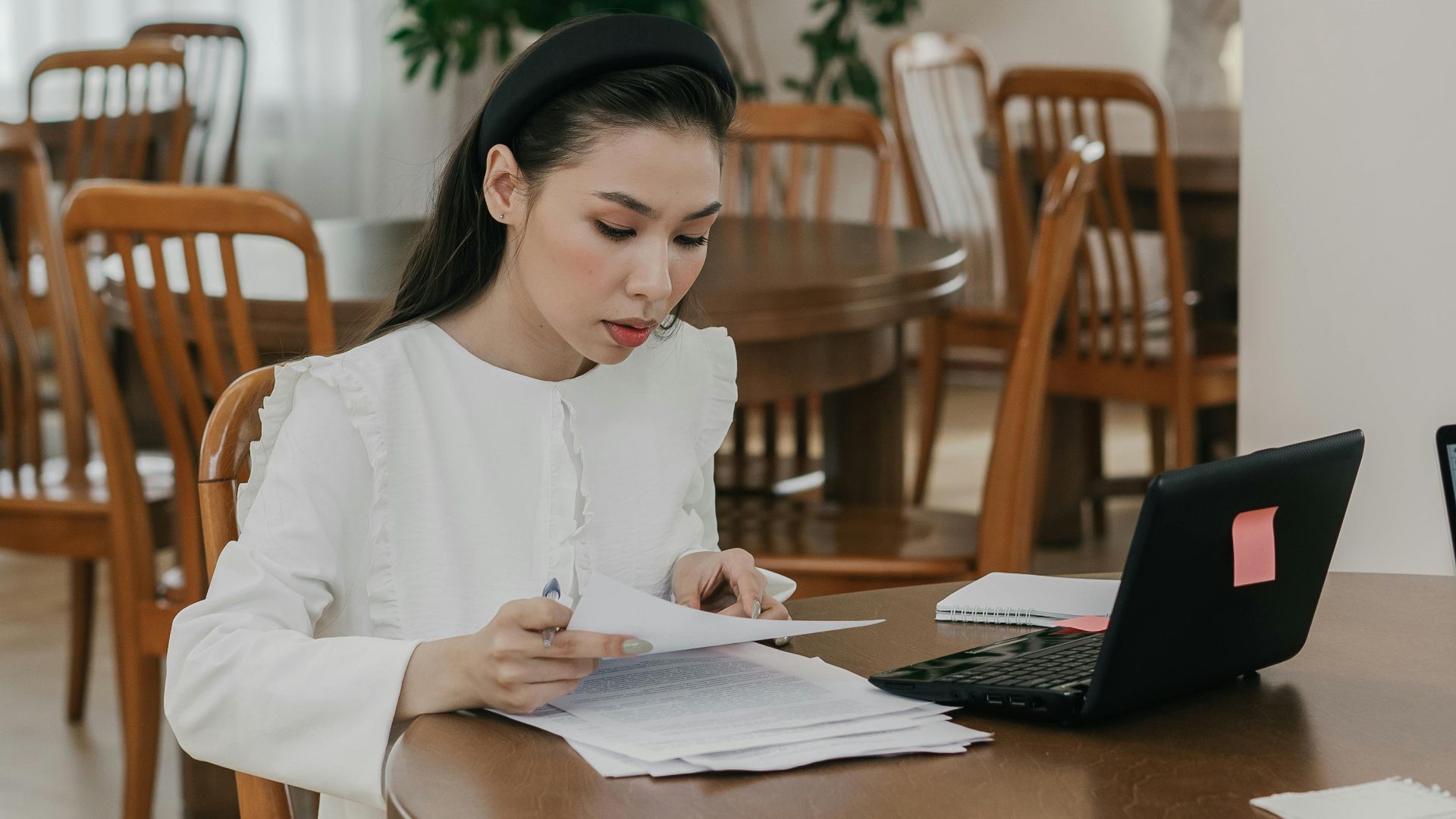 Focused woman studying with papers and laptop in a quiet library setting, surrounded by wooden tables and chairs.